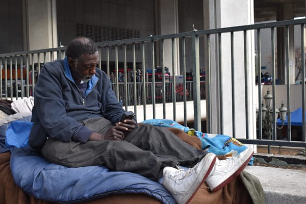 A dark skinned Black man wearing a navy blue jacket and gray jeans with white sneakers sits on a sack of blankets. 
